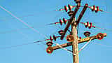 A wooden utility pole with ceramic insulators and a high voltage sign stands against a bright blue sky.
