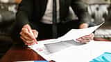 A professional in a suit uses a red marker to review and highlight details on a printed financial document.