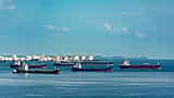 Several oil tankers wait on the water in front of a coastal fuel storage terminal with large white tanks.