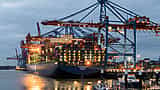 A massive, fully loaded container ship illuminated by warm lights is docked under large cranes at a bustling commercial terminal at dusk.