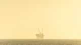 An offshore oil platform sits on the horizon under a golden, hazy sky.