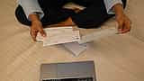 A person's hands hold printed checks over a desk with a laptop.