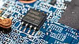 A black microchip is soldered onto a blue printed circuit board among other small electronic components.
