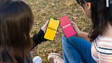 Two people sit outdoors holding smartphones that display the Bumble and Tinder dating apps.