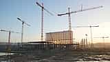 Multiple towering cranes stand over a massive building framework at a large-scale construction site at dusk.