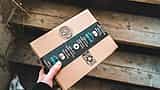 A hand holds a cardboard delivery box sealed with Amazon Prime tape over wooden steps.