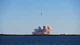 A silver rocket launches into a clear blue sky, leaving a large plume of smoke over a flat coastal landscape.