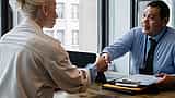 A smiling manager shakes hands with a candidate across a desk in a bright, modern office.