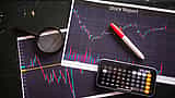 A printed stock report showing candlestick charts, resting on a dark surface alongside a magnifying glass, marker, and a calculator on a smartphone.
