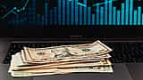 A stack of US dollar bills rests on a laptop keyboard with a glowing blue financial chart displayed on the screen.