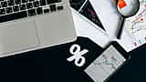A white percentage symbol rests on a dark desk surrounded by a laptop, a smartphone displaying market data, and printed financial charts.