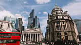 A red double-decker bus passes historic classical buildings and modern glass skyscrapers in the City of London's financial district.