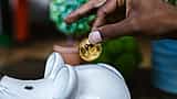 A hand places a gold physical Bitcoin token into the slot of a white ceramic piggy bank.