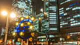 The illuminated Euro sculpture stands in front of office buildings at night in Frankfurt, Germany.