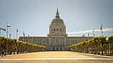 The grand, dome-topped San Francisco City Hall building viewed from a wide, sunlit plaza lined with trees.