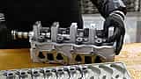 A worker wearing black gloves handles a newly machined metal engine component on a workbench.