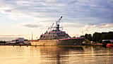 A modern U.S. Navy warship docked in a serene harbor bathed in golden sunset light.