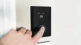 A close-up of a person's finger pressing the dial on a sleek, black digital smart thermostat mounted on a wall.