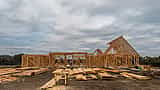The wooden frame of a large, unfinished house stands under a cloudy, overcast sky.