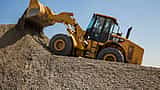 A yellow Caterpillar front loader carrying gravel up a steep mound under a clear blue sky.
