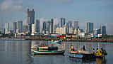Traditional fishing boats float in a harbor with modern skyscrapers under construction in the background.