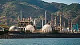 A large industrial oil refinery with spherical storage tanks and tall towers sits near a body of water.