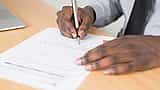 Close-up of a businessman in a grey shirt and tie signing a formal document with a fountain pen.