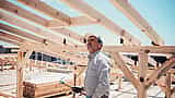 A construction worker holding a hammer stands beneath the exposed wooden roof trusses of a new home build.
