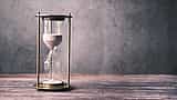 A brass hourglass filled with white sand sits on a wooden surface against a dark, mottled background.