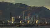 A large industrial oil refinery with storage tanks, piping, and smokestacks operating at the base of a hazy, rugged mountain range.