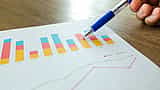 A person's hand holds a blue pen, pointing at a colorful printed financial bar chart resting on a wooden desk.