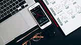 A smartphone displaying a downward-trending stock market chart sits on a desk next to a laptop, eyeglasses, and printed market analysis documents.
