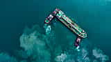 An aerial view of a large tanker ship being guided by two tugboats through bright turquoise water.
