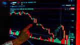 A person's finger points to a sharply falling red candlestick chart displayed on a glowing computer screen.