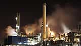 A sprawling industrial oil refinery illuminated at night, with smokestacks emitting thick plumes into the dark sky.