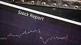 A document horizontally labeled Stock Report rests on a digital screen displaying fluctuating financial market charts.