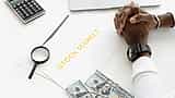 A person rests folded hands on a desk with stock market charts, a calculator, and scattered cash.