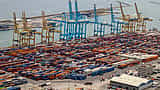 Thousands of brightly colored shipping containers sit stacked at a large coastal port with blue loading cranes.