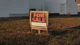 A real estate 'For Sale' sign stands in the front yard of a suburban home at sunset.