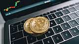 Two physical gold bitcoins rest on a laptop keyboard in front of a screen displaying a financial candlestick chart.