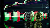 Two physical gold Bitcoin tokens stand on a reflective desk in front of a glowing monitor displaying a cryptocurrency trading chart.