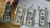 A person's hand counts US dollar bills over a desk covered in financial performance charts.
