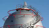 A large, spherical industrial natural gas storage tank with a metal staircase winding around it against a blue sky.