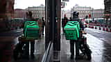 An Uber Eats courier wearing a green delivery backpack stands on a wet city street, reflected in a nearby window.