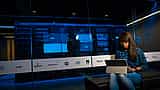 A woman works on a laptop in a server room behind a glass wall displaying the logos of various technology companies.