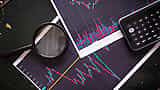 A magnifying glass and a smartphone calculator rest on printed stock market charts.