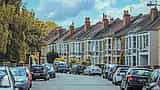 A row of classic British terraced houses stretches down a residential street beneath a partly cloudy sky.