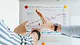 Two people pointing at a printed financial market chart pinned to a whiteboard.