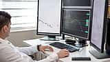 A financial professional analyzes market charts on multiple computer monitors in an office.