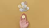 A hand holding a physical cryptocurrency coin above a scattered pile of digital currency coins on a bright yellow background.